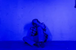 A person sitting against a wall in a blue-lit room, expressing solitude and contemplation.
